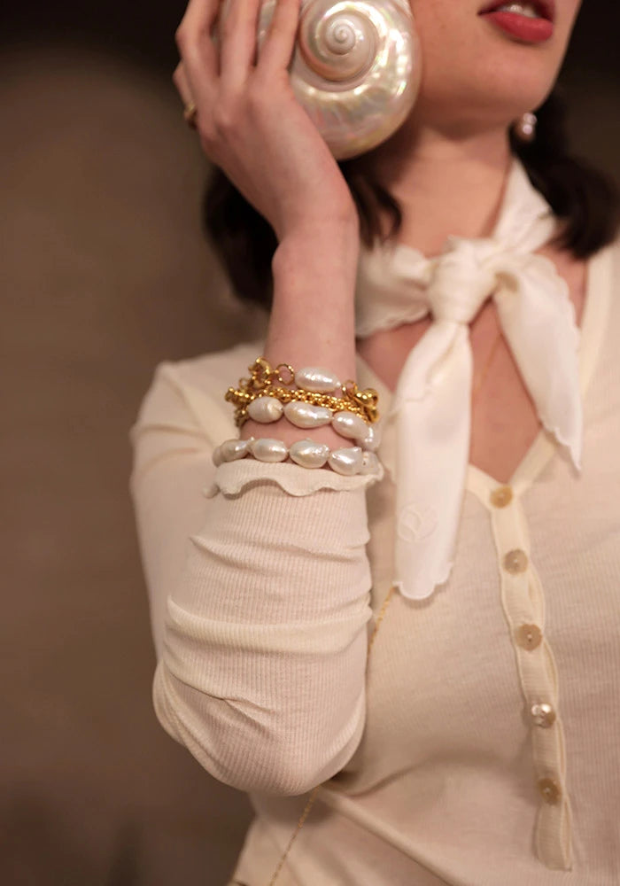 Une femme porte un gros coquillage nacré à l'oreille. À son poignet, quatre gros bracelets variés, composés de grosses perles baroques irrégulières et de chaînes en laiton brillant. Elle arbore un tee-shirt en côtes de coton biologique GOTS écru sans teinture, boutonné avec de petites fleurs en nacre, et un foulard en soie écru autour du cou. Foulard en soie made in France. Bracelets made in France. Bracelet en perles baroques et laiton. T-shirt femme made in France. Cadeaux pour femme made in France.