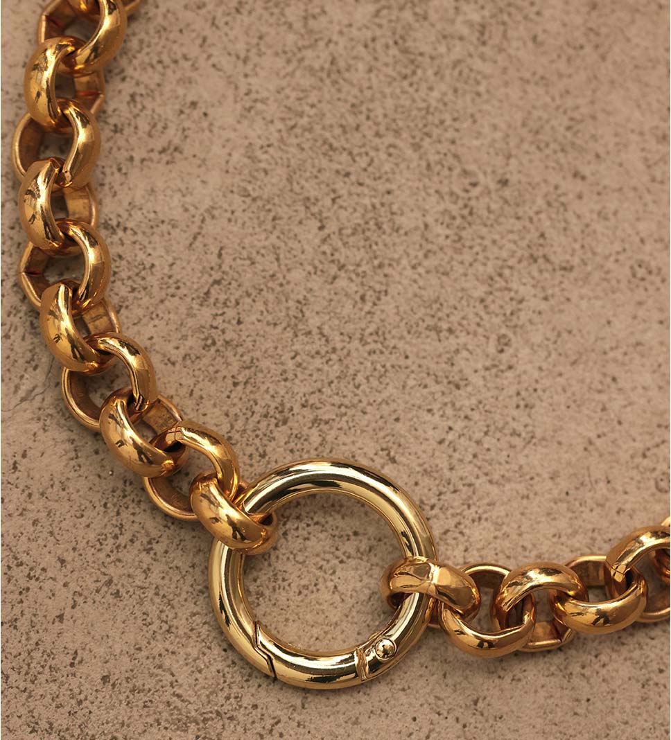 Gros plan sur un collier en laiton, composé de grosses mailles rondes et d'un mousqueton également rond, d'une teinte dorée claire, déposé sur un sol en béton ciré de couleur sable, composants italiens, assemblage à Paris.