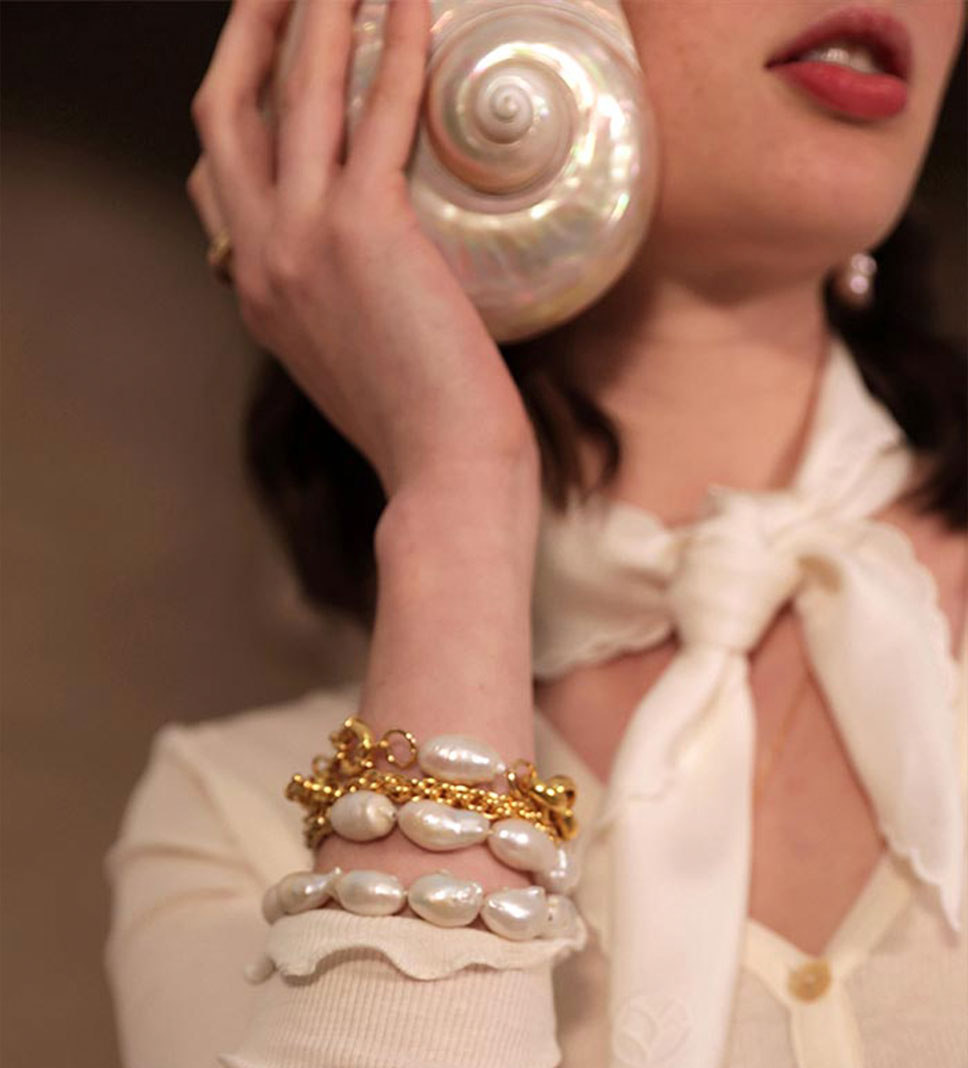 Une femme porte un gros coquillage nacré à l'oreille. À son poignet, quatre gros bracelets variés, composés de grosses perles baroques irrégulières et de chaînes en laiton brillant. Elle arbore un tee-shirt en côtes de coton biologique GOTS écru sans teinture, boutonné avec de petites fleurs en nacre, et un foulard en soie écru autour du cou.