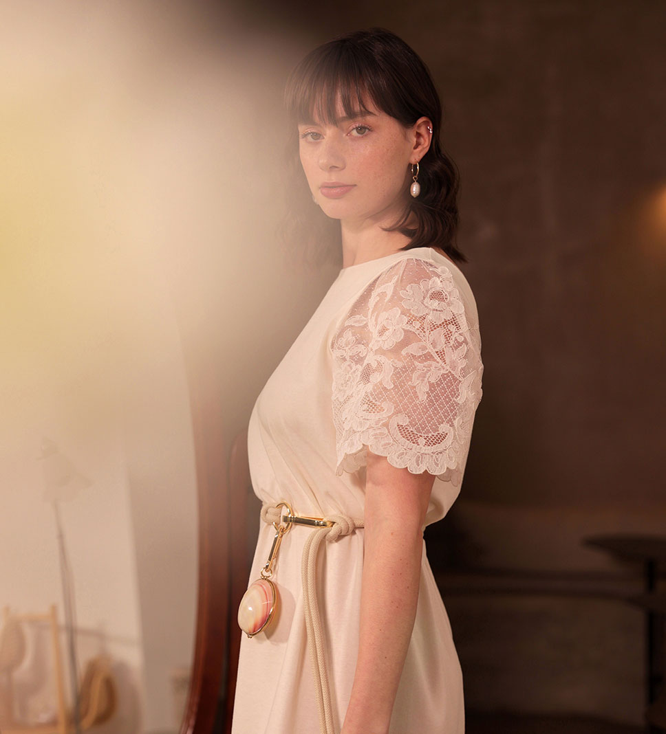 Dans un élégant rendu artistique flouté, une femme est vêtue d'une robe écrue aux manches en dentelle de Lyon à motif floral, sa taille est soulignée par une ceinture en corde écrue fermée par un mousqueton XXL où est accroché un petit sac en coquillage et laiton. l'ensemble est réalisé en France.