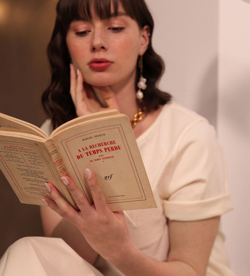Une femme brune lit le roman 'À la Recherche du Temps Perdu' de Marcel Proust. Elle porte un tee-shirt en coton biologique certifié Gots écru avec une poche en forme de tulipe, manches retroussées. Autour de son cou, un collier en gros maillons en laiton brillant, et elle arbore des pendants d'oreilles avec deux grosses perles baroques de forme irrégulière. Le tout est fabriqué en France.