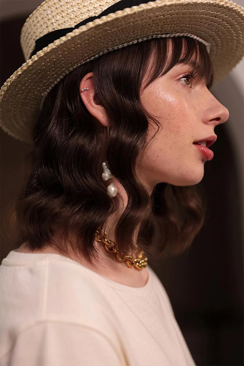 Femme lumineuse, canotier noir, coupe ondulée avec boucle d'oreille en perles baroques, collier laiton brillant, tee-shirt écrue en coton biologique certifié GOTS à encolure bateau. Son regard plein d'espoir vers l'avenir, sa tenur fabriquée en France.