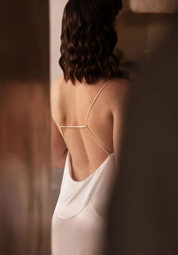 Une femme, vue de dos, porte une robe dos nu évoquant le style nuisette, confectionnée en France dans une soie biologique écrue sans teinture. Des bretelles graphiques et des découpes artistiques autour des hanches créent l'effet de pétales gracieux d'une fleur. Une ombre au premier plan suggère l'approche d'un homme vers elle.
